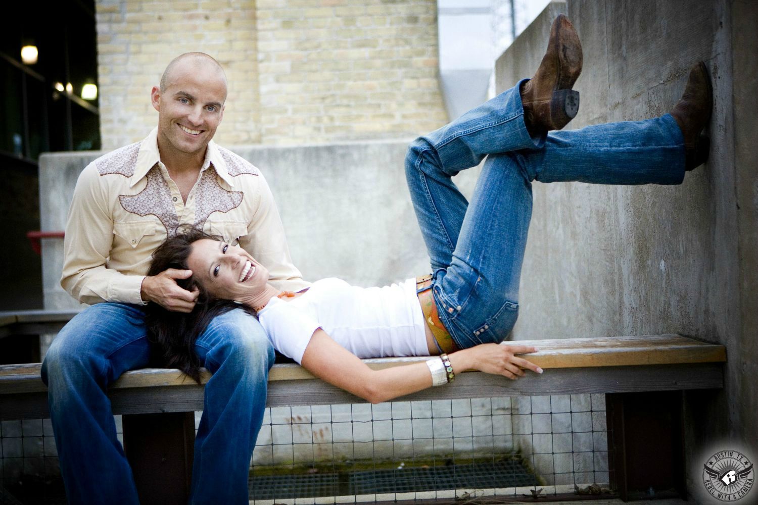Super skinny, hot  brunette gal in a tight white scoop neck shirt, blue jeans and brown cowboy boots lies on a bench with her head on the lap of a sitting  guy wearing a cowboy shirt and blue jeans with epic concrete and brick in the background of this fantastic engagement photo in SOCO in Austin.